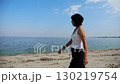 Happy adult woman going on sandy beach near shoreline at sunny day. Beautiful lady walking on seashore with calm waves at background. Concept of summer vacation or holiday. Slow motion 130219754