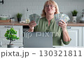 Mature blonde woman wearing eyeglasses celebrating winnings. Female holding dollar banknotes while sitting at kitchen with laptop, expressing joy and financial success. Makes fan and waves banknotes 130321811