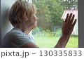 Elderly woman with gray hair and deep wrinkles on skin looks longingly out window at backyard. Hand touches glass, as if it is trying to reach outside world. Self isolation old ill people 130335833