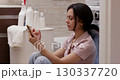 brunette woman using smartphone while laundry is in washing machine, woman sitting on the floor near washing machine in bathroom 130337720