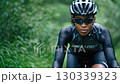 A focused cyclist wearing a sleek helmet and sunglasses, navigating a scenic path surrounded by lush greenery and nature's beauty. 130339323