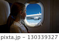 A young girl gazes out the airplane window, captivated by the stunning view of the clear blue sky and the aircraft wing in flight. 130339327