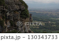 Two climbers on via ferrata route navigating exposed cliffside cables with panoramic valley and farmland views in background 130343733