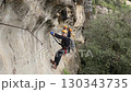 Two climbers navigating exposed via ferrata cliff traverse secured to steel cable above forested gorge in mountainous terrain 130343735