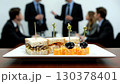 Group of professionals gathers in meeting room for networking. Plates of sandwiches and cheese cubes are arranged on table. Casual conversations and drinks enhance atmosphere 130378401