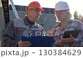 Construction professionals wearing safety helmet collaborating while reviewing blueprints and discussing project details at outdoor building development site. Foreman and architect showcasing teamwork 130384629