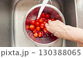 A woman washes fresh cherry tomatoes under running water in a metal colander, preparing vegetables for cooking a healthy meal in a clean kitchen environment 130388065