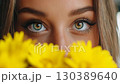 Young woman with striking eyes peeking through vibrant yellow flowers in a close-up shot during daylight 130389640