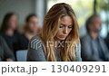 A pensive woman with long hair gazes thoughtfully during a group discussion in a modern, softly lit room. 130409291