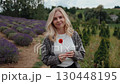 Blonde woman wearing casual clothes, holding poppy flower in her hands, going in lavender field, enjoying moment and peaceful expression. Blonde smiling Caucasian woman in her 30s walking in field 130448195
