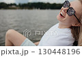 Blond woman enjoying sunny lake day, pedaling leisurely on water with sunglasses, laughing while exploring peaceful surroundings during summer vacation. Blond caucasian woman pedaling kayak, laughing 130448196
