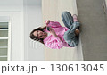 A young woman in jeans and a pink shirt sits cross-legged on the pavement, holding her phone. The shaky live-camera style adds realism, showing relaxation, music, and everyday urban lifestyle. 130613045