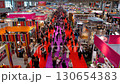 Aerial view of a bustling trade show, teeming with attendees navigating through numerous exhibitor booths in a busy indoor commercial fair. 130654383