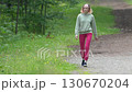 Full length view of female fitness instructor walking along footpath in park during summer sports training. Slim athletic sportswoman dressed in sportswear - light green hoodie, red joggers. Zoom out Full length view of female fitness instructor walking along footpath in park during summer sports training. Slim athletic sportswoman dressed in sportswear - light green hoodie, red joggers. Zoom out 130670204