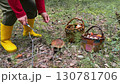 Individual wearing yellow rubber boots is foraging for mushrooms in a lush forest, surrounded by baskets filled  130781706