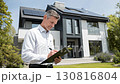 Professional man in a white shirt, inspecting a modern home with solar panels, taking notes on a clipboard, showcasing meticulous attention to detail in property assessment 130816804