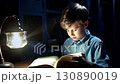 Young man reading oversized book, soft desk lamp casting warm glow, concentrating intently while turning pages during peaceful evening study session 130890019