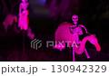 Close-up Halloween yard decoration shows a small skeleton riding a pink lawn flamingo under purple light. The spooky outdoor scene glows at night with neon bokeh and shallow depth of field. 130942329