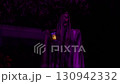 Close-up Halloween yard decoration shows a hooded ghost holding a lantern in purple light at night. Spooky outdoor scene with draped fabric, skull face, and a glowing lamp. 130942332