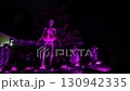 Front yard Halloween display with a giant skeleton, pink flamingos, ghosts, and tombstones glowing in purple light at night. Spooky suburban house scene shows neon lights, eerie decorations, and 130942335