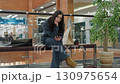 Girl in dark outfit seated on bench inside bright shopping mall browses phone calmly, shopping bags on both sides, surrounded by glass railing, potted tree and modern retail background 130975654