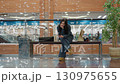 Young woman dressed in dark outfit seated on bench inside brightly lit shopping mall, browsing phone with relaxed posture, paper shopping bags beside her, near glass railing and potted green plant 130975655