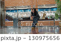 Young woman in dark outfit seated on bench inside shopping mall browses her phone with relaxed expression, shopping bags beside her on bench, surrounded by glass railing and potted plant 130975656