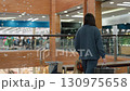 Rear view of shopper walking toward bench inside brightly lit shopping mall, holding black and brown paper bags, preparing to sit in relaxed posture as crowd shops in background behind glass railing 130975658