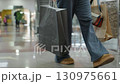 Shopper walking inside mall corridor with shopping bags in both hands, brown shoes moving across polished floor while approaching bench, with people and descending escalator in background 130975661