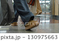 Low angle close up of shopper legs in brown shoes walking across polished tile floor of modern shopping mall, carrying shopping bags while heading toward bench near escalator in busy retail setting 130975663
