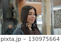 Young woman with long hair walks through shopping mall with eyes closed and soft smile, preparing to sit on bench, surrounded by tiled walls, warm ceiling lights, and blurred background of escalator 130975664