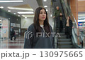 Young female shopper stands calmly in modern shopping mall after shopping, looking thoughtful while people descend escalator behind her, surrounded by shops, plants, and bright overhead lights 130975665