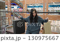 Young woman in loose sweatshirt walks inside modern shopping mall after completing shopping, looking around while waiting for friend, surrounded by glass railing and colorful background 130975667