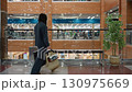 Young girl in dark outfit finishes shopping inside modern mall, drops shopping bag on bench, sits down, crosses leg beside indoor plant and glass railing with view of clothing store behind her 130975669