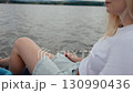 Young blonde woman wearing white t shirt and light blue denim shorts enjoys relaxing pedal boat ride on calm lake, surrounded by beauty of nature. Blonde woman relaxing on pedal boat. Recreation mood 130990436