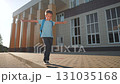 Playful boy walking on pavement balancing arms. Student walking outdoors after school. Schoolboy backpack steps carefully. Child balancing near building. Boy walking sidewalk. Schoolboy playful walk. 131035168