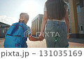 Kid with backpack holding woman hand walks near school. Boy walks outdoors with mother. Child walking beside woman slowly. Backpack on boy. School in background. Outdoor scene with kid walking. 131035169