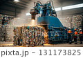 A hydraulic baler efficiently compresses mixed recyclable materials into compact cubes. Workers in protective gear monitor the process in a busy industrial setting with sparks and dust. 131173825