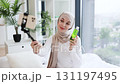 Young Muslim female blogger recording video with smartphone selfie stick promoting facial moisturizer. Indoor setting with natural lighting and green plants. 131197495