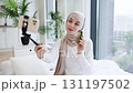 Young Muslim woman in beige hijab holding smartphone on tripod sharing skincare tips using avocado. She discusses natural beauty methods while creating engaging and insightful content in a cozy home 131197502