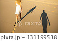 A woman strolls along a serene beach, leaving footprints in the sand. Unbeknownst to her, a shadow indicating an attacker lurks nearby, creating an atmosphere of tension. 131199538