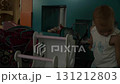 A baby girl with short blond hair takes a large doll out of a wooden white stroller, continuing to take out all sorts of things in a row, focused and busy, at a play center 131212803