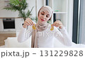 Young Muslim woman focused on skincare routine using gold patches on eye area, showcasing beauty and self-care. Relaxing indoor setting, wearing beige hijab and white top, maintaining wellness. 131229828