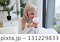 Smiling adult Muslim woman wearing hijab applying moisturizer cream on facial skin indoors. Promotes skincare, self-care, beauty regime, and wellness. 131229833