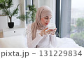 Young adult Muslim woman in hijab skincare routine at home with facial cream. Expresses happiness and simple beauty self-care. Bright indoor atmosphere showcasing plant in stylish decor 131229834