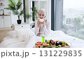 Smiling young Muslim woman wearing hijab applying moisturizing cream to face, sitting on bed in bright modern rooms, surrounded by skincare and fresh fruits showcasing self-care, skincare 131229835