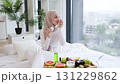 Young Muslim woman sitting on bed using cotton pads for skincare, surrounded by healthy breakfast including fruits and drinks, within a bright and cozy interior setting, promoting beauty and wellness. 131229862