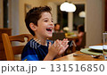 Happy caucasian boy child laughing and clapping hands while sitting at dining table in warm family kitchen during dinner time 131516850