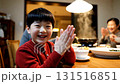 Happy Asian boy in red shirt waving and clapping hands at family dinner table in cozy restaurant with warm lighting 131516851