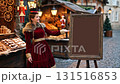 European woman in vintage burgundy dress presenting blank chalkboard sign at festive Christmas market with traditional wooden stalls and holiday decorations 131516853